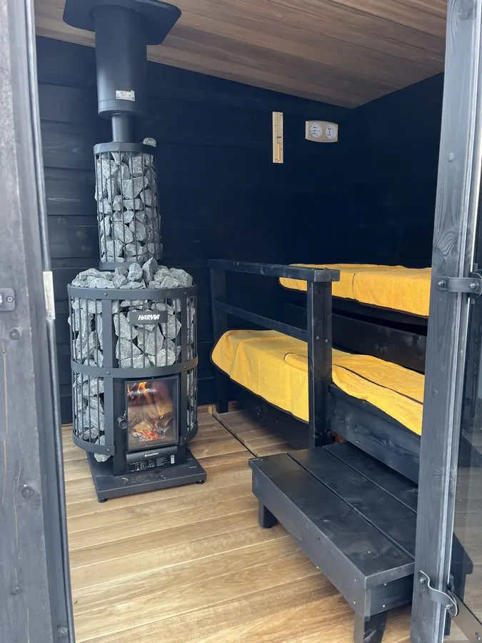 サウナ小屋の内部。石を積んだハルビアの薪ヒーターに炎が入り、黒い壁と、黄色いタオルを敷いた二段のベンチ