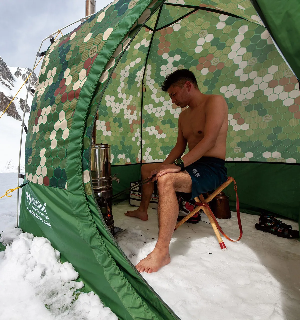 雪山のフィールドでテントサウナを楽しむ男性、Mobibaテントの中で椅子に座り薪ストーブの前でくつろぐ
