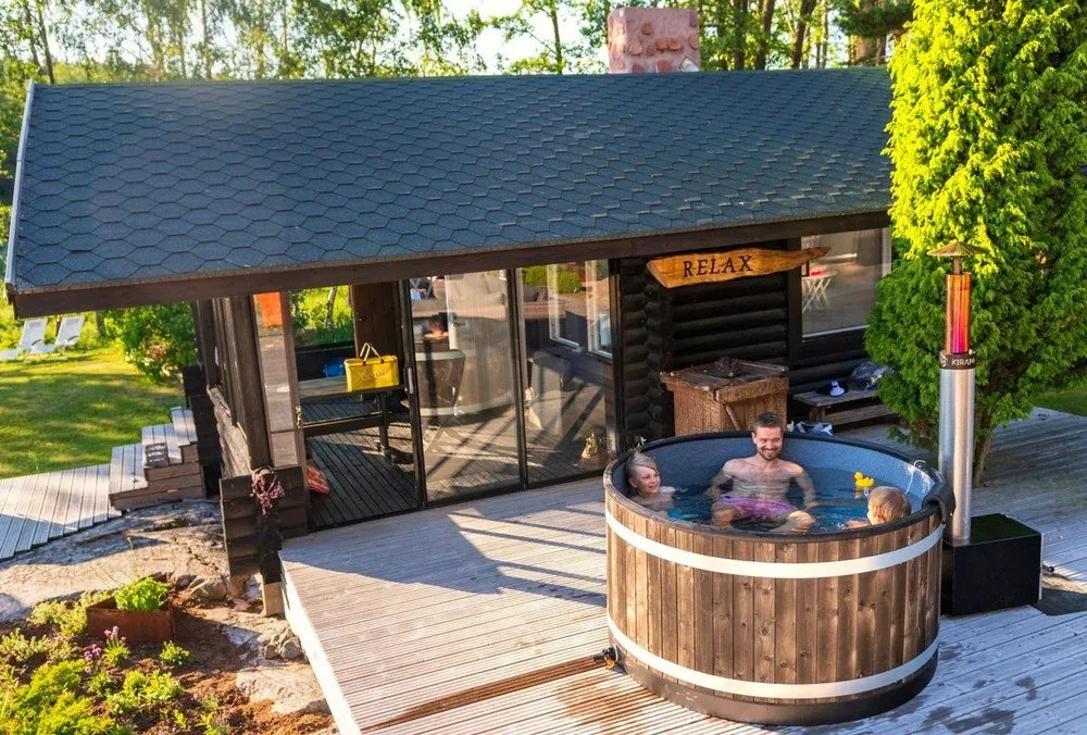 黒い山小屋の前の木のデッキに置かれたホットタブ。3人が湯に浸かり、右手に銀色の煙突。小屋には RELAX の看板（写真：HARVIA）